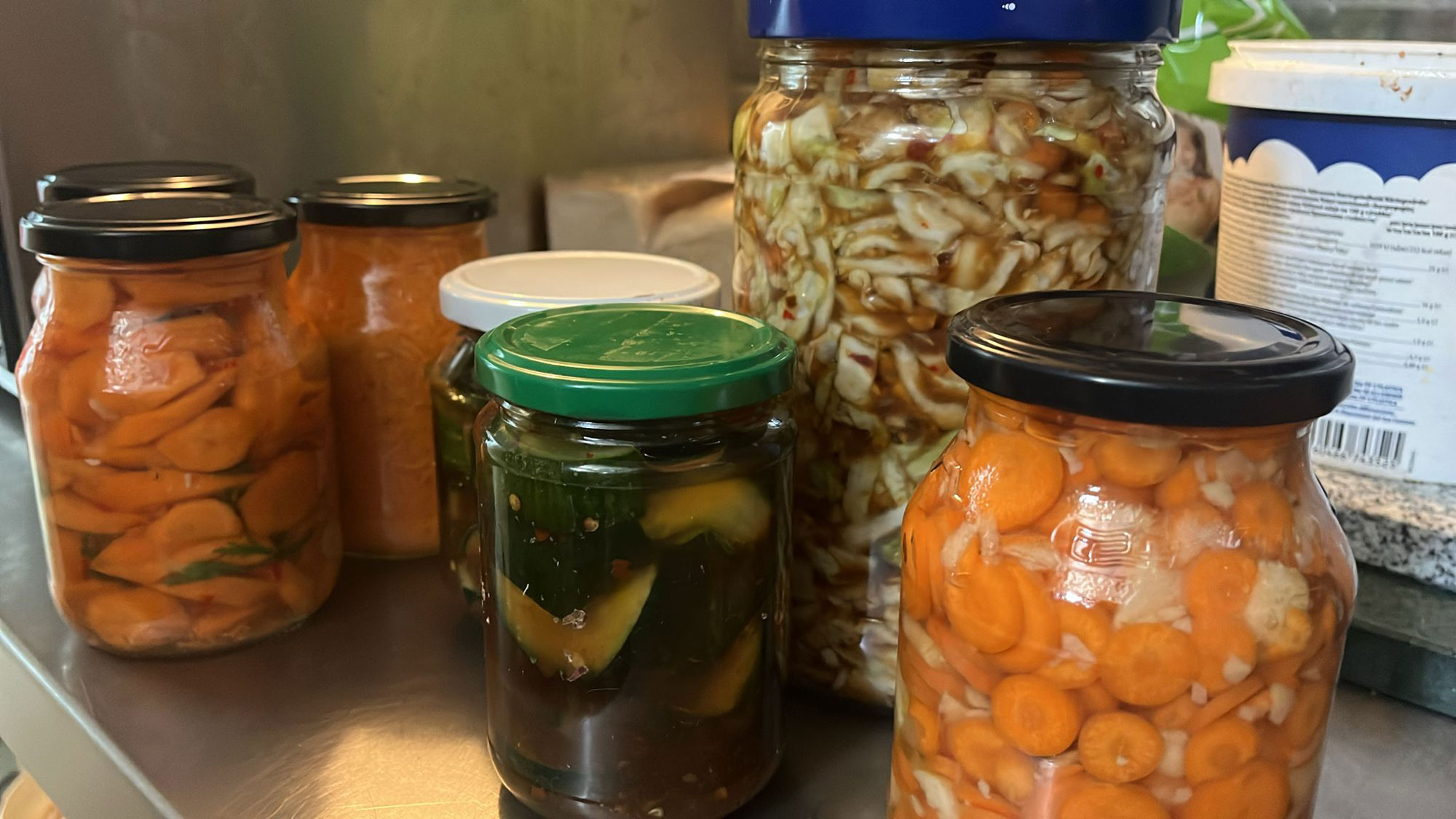 Mehrere Gläser mit fermentiertem Gemüse stehen auf einer Edelstahl-Arbeitsfläche. Zu sehen sind unterschiedlich befüllte Gläser mit Karotten, Gurken, Weißkohl und weiteren eingelegten Gemüsemischungen. Das Bild zeigt Ergebnisse aus dem Fermentations-Workshop des Schmidt’s Restaurant Dresden, bei dem Mitarbeitende traditionelle Techniken zur nachhaltigen Haltbarmachung saisonaler Zutaten ausprobieren.
