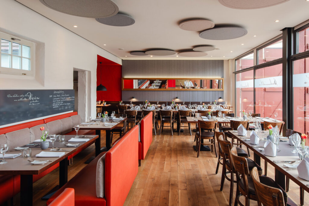 Der Gastraum des Schmidt’s Restaurant Dresden zeigt einen großzügigen, hellen Dining-Bereich mit eingedeckten Tischen, weißen Servietten, frischen Tulpen und polierten Weingläsern. Große Fenster und warme Holzböden prägen die moderne Bistro Atmosphäre im Hellerau Restaurant. Die Kombination aus roten Akzenten, langen Sitzbänken und stilvollen Leuchten vermittelt den Charakter eines Fine Dining Restaurants mit regionaler, saisonaler und kreativer Küche in Dresden.