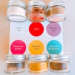 Sechs kleine Glasdosen der „Schmidt’s Gewürzbox 1“ stehen in zwei Reihen auf einer Karte mit farbigen Labels. Enthalten sind: Vanillesalz, Chili, Räuchersalz, Szechuanpfeffer, Schmidt’s Curry und Schmidt’s Wildgewürz – eine ausgewählte Gewürzmischung aus der Küche des Schmidt’s Restaurant Dresden.