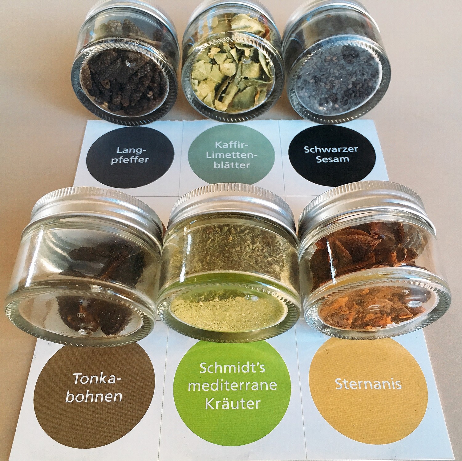 Sechs kleine Glasdosen mit Gewürzen aus der Schmidt’s Gewürzbox 2 stehen in zwei Reihen auf einer Karte mit farbigen Beschriftungen. Enthalten sind Langpfeffer, Kaffir-Limettenblätter, Schwarzer Sesam, Tonkabohnen, Schmidt’s mediterrane Kräuter und Sternanis. Eine hochwertige Auswahl – typisch für die Küche des Schmidt’s Restaurant Dresden.