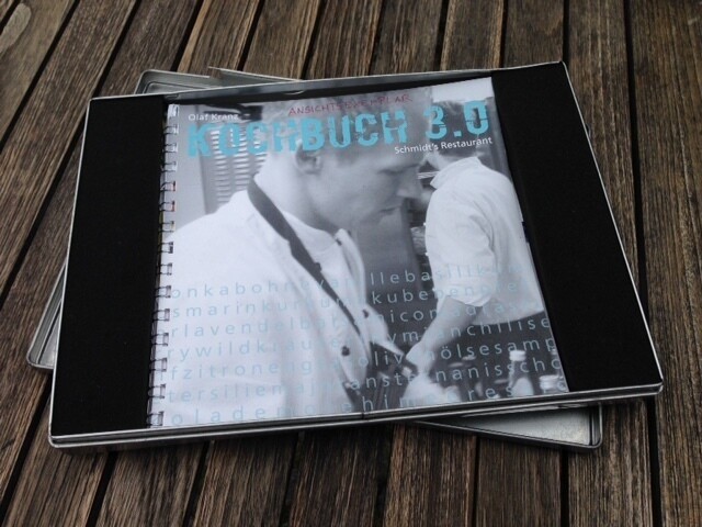 Eine flache, rechteckige Geschenkbox aus Metall liegt auf einem Holztisch. Darauf abgebildet ist ein Kochbuch mit einem schwarz-weißen Titelbild, das einen Koch in der Restaurantküche zeigt. Die Box gehört zum Sortiment des Schmidt’s Restaurant Dresden und dient als stilvolle Verpackung, zum Beispiel für das Kochbuch von Koch Olaf Kranz.