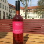 Eine Flasche „Schmetterlinge im Bauch“ Roséwein (feinherb) vom Weingut Kiefer aus Baden steht auf einem Holztisch im Innenhof von Dresden-Hellerau; sonnige Produktaufnahme für den Shop von Schmidt’s Restaurant Dresden.