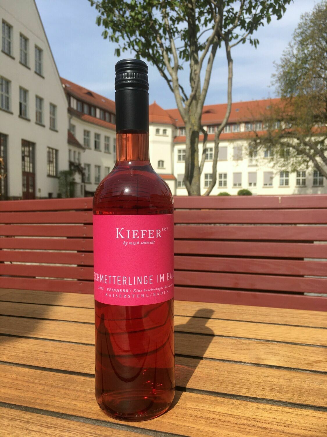 Eine Flasche „Schmetterlinge im Bauch“ Roséwein (feinherb) vom Weingut Kiefer aus Baden steht auf einem Holztisch im Innenhof von Dresden-Hellerau; sonnige Produktaufnahme für den Shop von Schmidt’s Restaurant Dresden.
