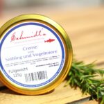 Ein Glas mit goldenem Schraubdeckel und dem Etikett ‚Schmidt’s – Creme von Saibling und Vogelmiere‘ steht auf einem Holztisch; daneben liegt ein Zweig frischer Kräuter. Ein hochwertiges Feinkostprodukt aus dem Shop des Slow-Food-Restaurants Schmidt’s Dresden.