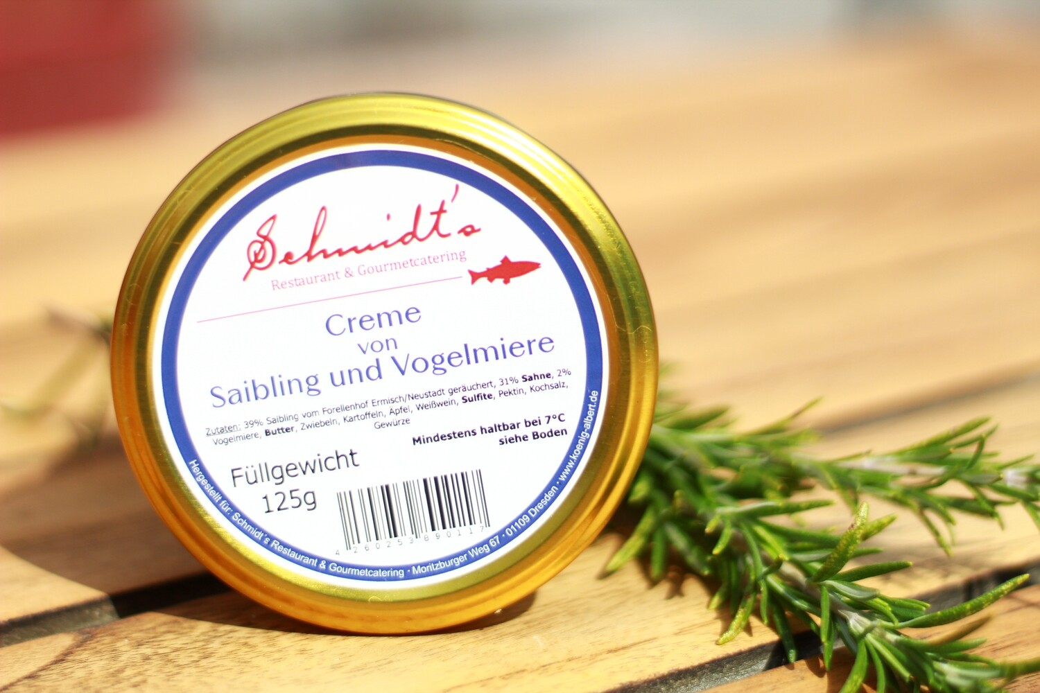 Ein Glas mit goldenem Schraubdeckel und dem Etikett ‚Schmidt’s – Creme von Saibling und Vogelmiere‘ steht auf einem Holztisch; daneben liegt ein Zweig frischer Kräuter. Ein hochwertiges Feinkostprodukt aus dem Shop des Slow-Food-Restaurants Schmidt’s Dresden.
