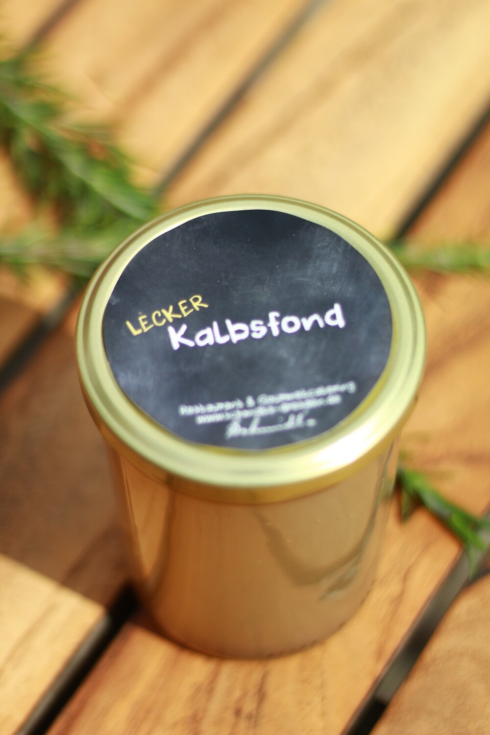 Ein Glas mit goldfarbenem Deckel und schwarzem Etikett mit der Aufschrift ‚Lecker Kalbsfond‘ steht auf einem Holztisch. Daneben liegt ein Zweig frischer Kräuter – ein Produkt aus dem Shop des Schmidt’s Restaurant Dresden.“