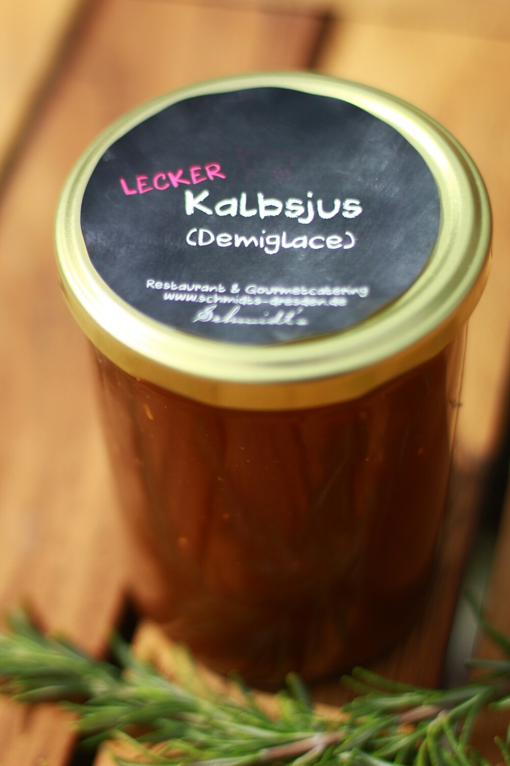 Ein Glas „Lecker Kalbsjus (Demiglace)“ aus dem Shop des Schmidt’s Restaurant Dresden steht auf einer Holzoberfläche. Der goldfarbene Deckel trägt ein schwarzes Etikett mit handschriftlicher Beschriftung; davor liegt ein Zweig frischer Rosmarin.