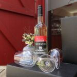 Eine Flasche Schmidt’s Lecker Weißwein vom Weingut Drei Herren steht vor einer roten Holztür; daneben liegen zwei elegante Weingläser sowie ein kleiner Strauß aus Wiesenblumen – stilvoll arrangiert als Genuss- und Produktpräsentation für den Shop im Schmidt’s Restaurant Dresden.