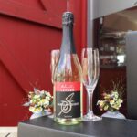 Eine Flasche Schmidt’s Sekt trocken aus dem Rheingau steht vor einer roten Holztür; daneben liegen zwei elegante Sektgläser sowie ein kleiner Strauß aus Wiesenblumen – stilvoll arrangiert als Genuss- und Produktpräsentation für den Shop im Schmidt’s Restaurant Dresden.