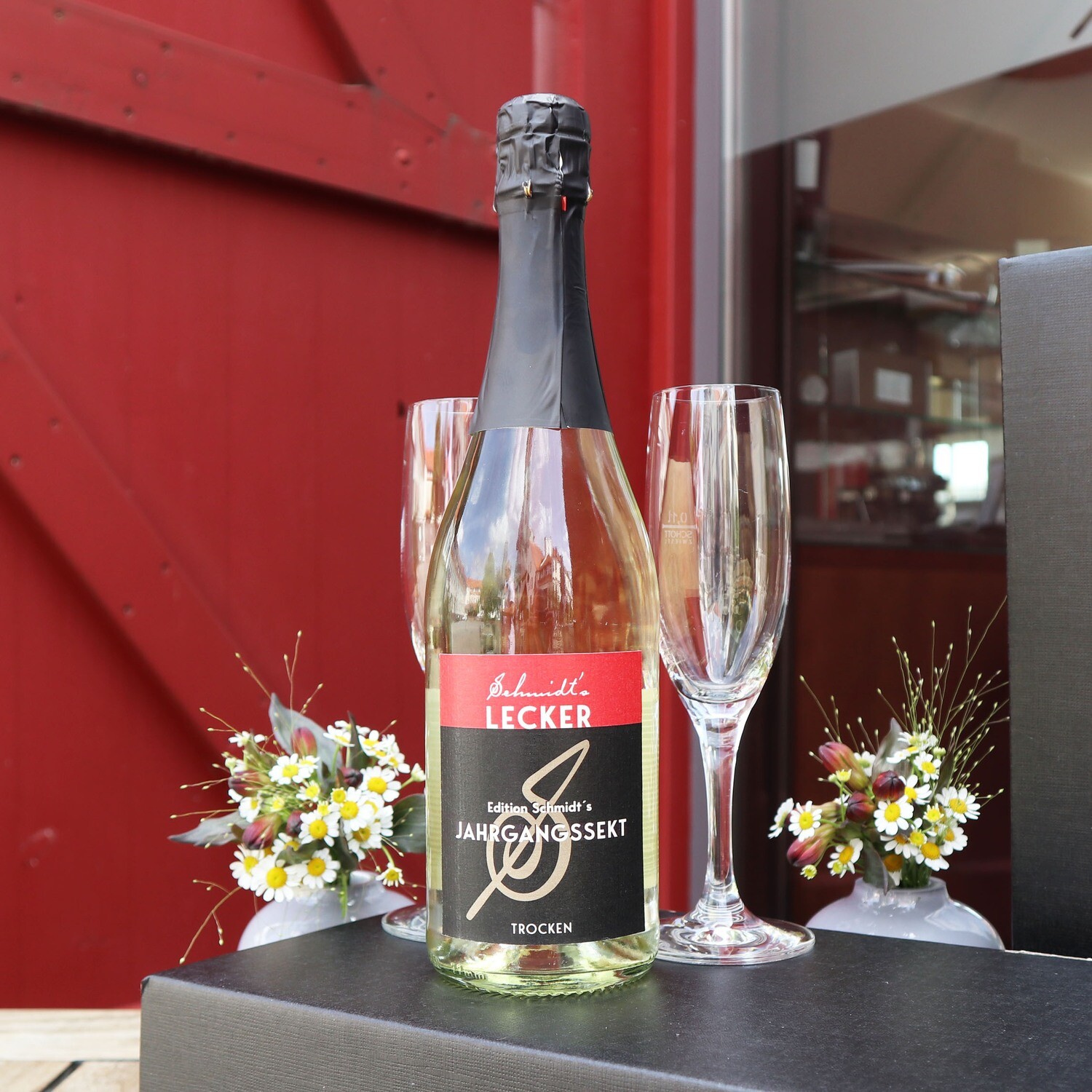 Eine Flasche Schmidt’s Sekt trocken aus dem Rheingau steht vor einer roten Holztür; daneben liegen zwei elegante Sektgläser sowie ein kleiner Strauß aus Wiesenblumen – stilvoll arrangiert als Genuss- und Produktpräsentation für den Shop im Schmidt’s Restaurant Dresden.