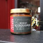 Ein Glas „Schmidt’s Lecker – Boeuf Bourguignon“ aus regionaler und nachhaltiger Küche steht vor rotem Hintergrund; das dunkle Etikett zeigt eine Rinderzeichnung, daneben ein kleines Blumenarrangement. Ein Produkt aus dem Shop des Schmidt’s Restaurant Dresden.