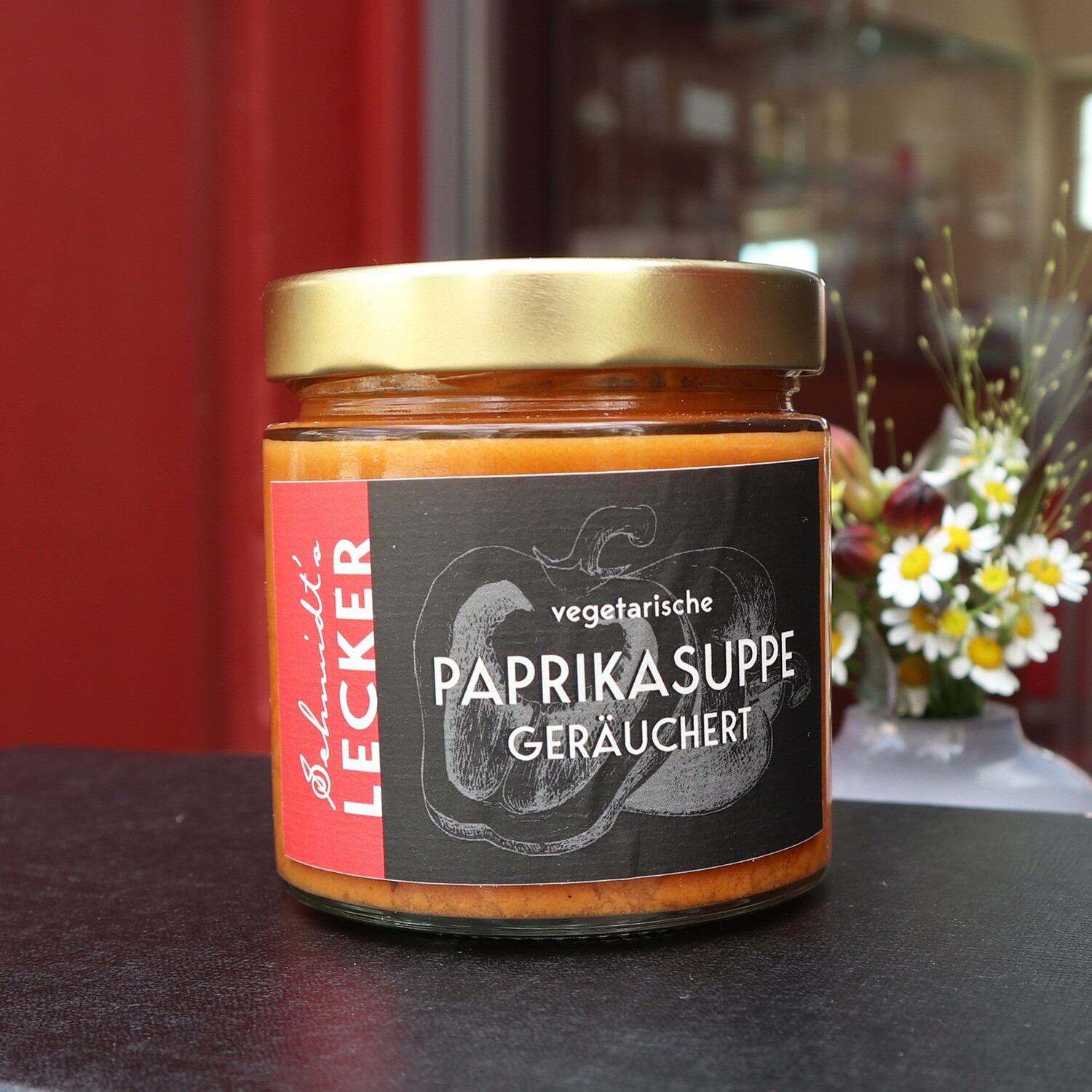 Ein Glas der „Schmidt’s Lecker – vegetarische Paprikasuppe geräuchert“ steht neben einem kleinen Blumenarrangement. Das Etikett zeigt eine gezeichnete Paprika und den Produktnamen. Die Suppe stammt aus dem Shop der kreativen Küche des Schmidt’s Restaurant Dresden.