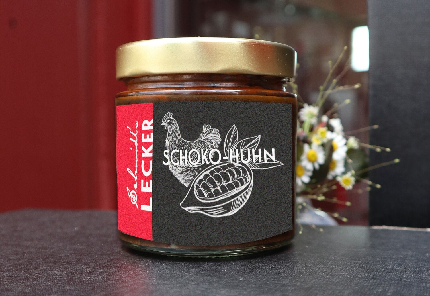Ein Glas „Schmidt’s Lecker – Schoko-Huhn“ aus dem Shop des Schmidt’s Restaurant Dresden. Das Glas steht vor rotem Hintergrund; das schwarze Etikett zeigt eine gezeichnete Kakaoschote und ein Huhn – regionale, kreative Küche im Glas.