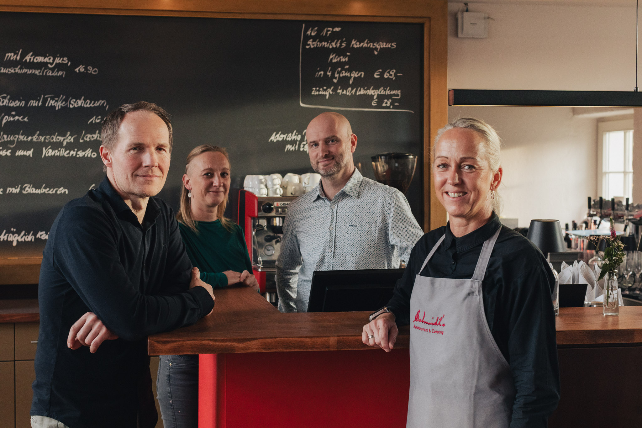 Das Team des Schmidt’s Restaurant Dresden steht an der Theke des modernen Slow Food und Fine Dining Restaurants in Hellerau. Vor einer großen Kreidetafel mit handgeschriebenem Menü der kreativ sächsischen Küche posieren vier Mitarbeitende, darunter Koch Olaf Kranz, in entspannter Bistroatmosphäre.