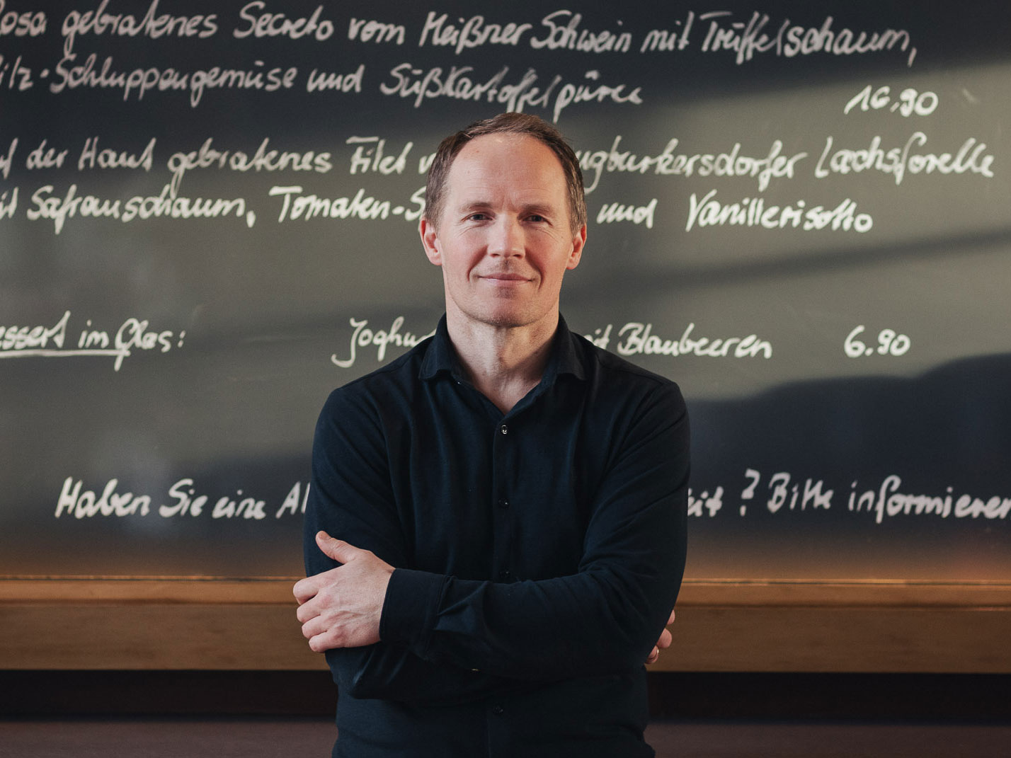 Koch Olaf Kranz lächelt freundlich in die Kamera und steht im Schmidt’s Fine-Dining-Restaurant in Dresden; vor einer großen, handbeschriebenen Tafel mit saisonalen Gerichten.