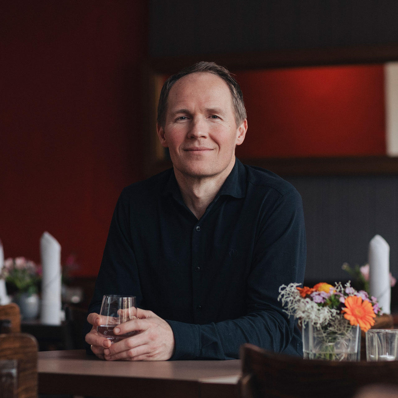 Koch Olaf Kranz des Schmidt’s Restaurant Dresden sitzt entspannt im Gastraum und lächelt freundlich in die Kamera. Er hält ein Glas in den Händen, auf dem Tisch stehen kleine Vasen mit frischen Blumen. Die warme, moderne Bistro-Atmosphäre des Slow-Food-Restaurants in Hellerau ist im Hintergrund erkennbar.