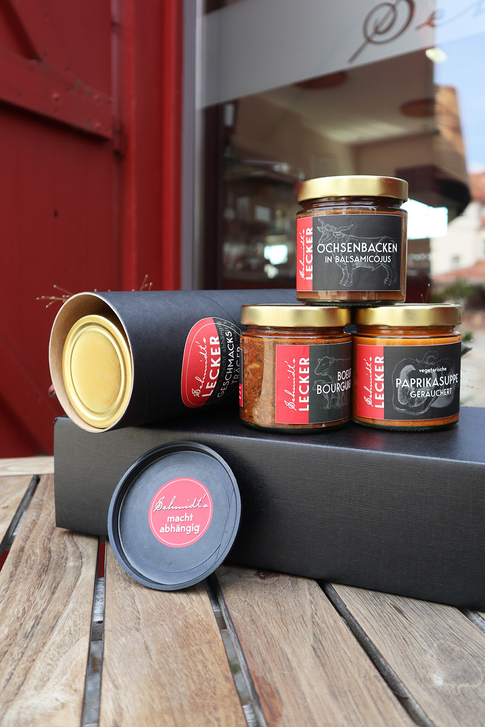 Schmidt’s Genussbox ‚Genuss Hoch Drei‘: Drei regionale Feinkost-Gläser aus dem Schmidt’s Restaurant Dresden – Ochsenbacken in Balsamicojus, Boeuf Bourguignon und geräucherte Paprikasuppe – stilvoll arrangiert mit schwarzer Geschenkbox und Runddose auf einem Holztisch vor dem Restaurant in Hellerau, Dresden.