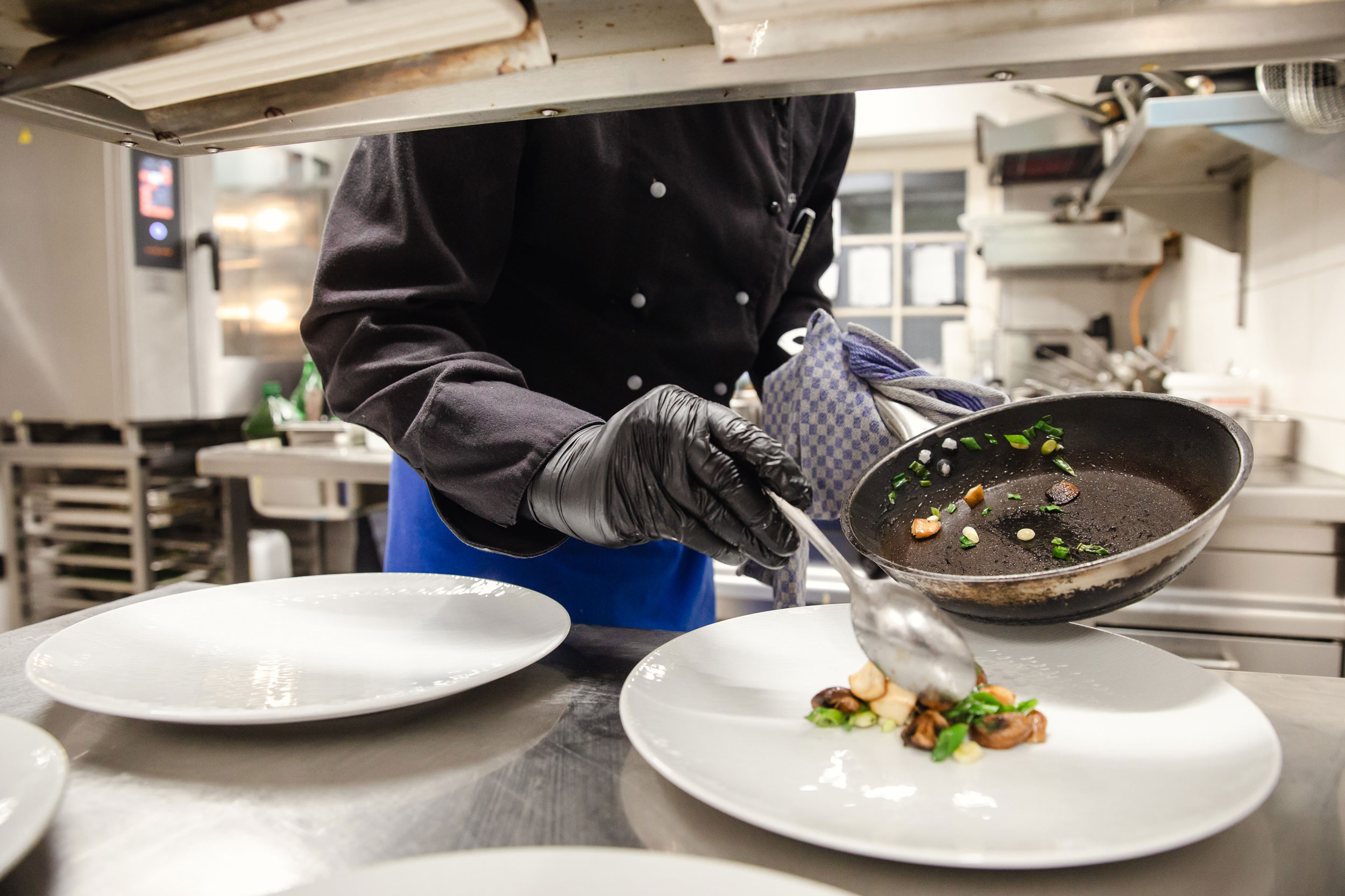 Ein Koch richtet ein Gericht auf einem großen weißen Teller an. Mit einer Pfanne in der einen Hand und einem Löffel in der anderen platziert er sautiertes Gemüse und Kräuter präzise auf dem Teller. Die Szene betont die kreative, saisonale und regional inspirierte Küche des Slow Food Restaurants Schmidt’s Dresden.