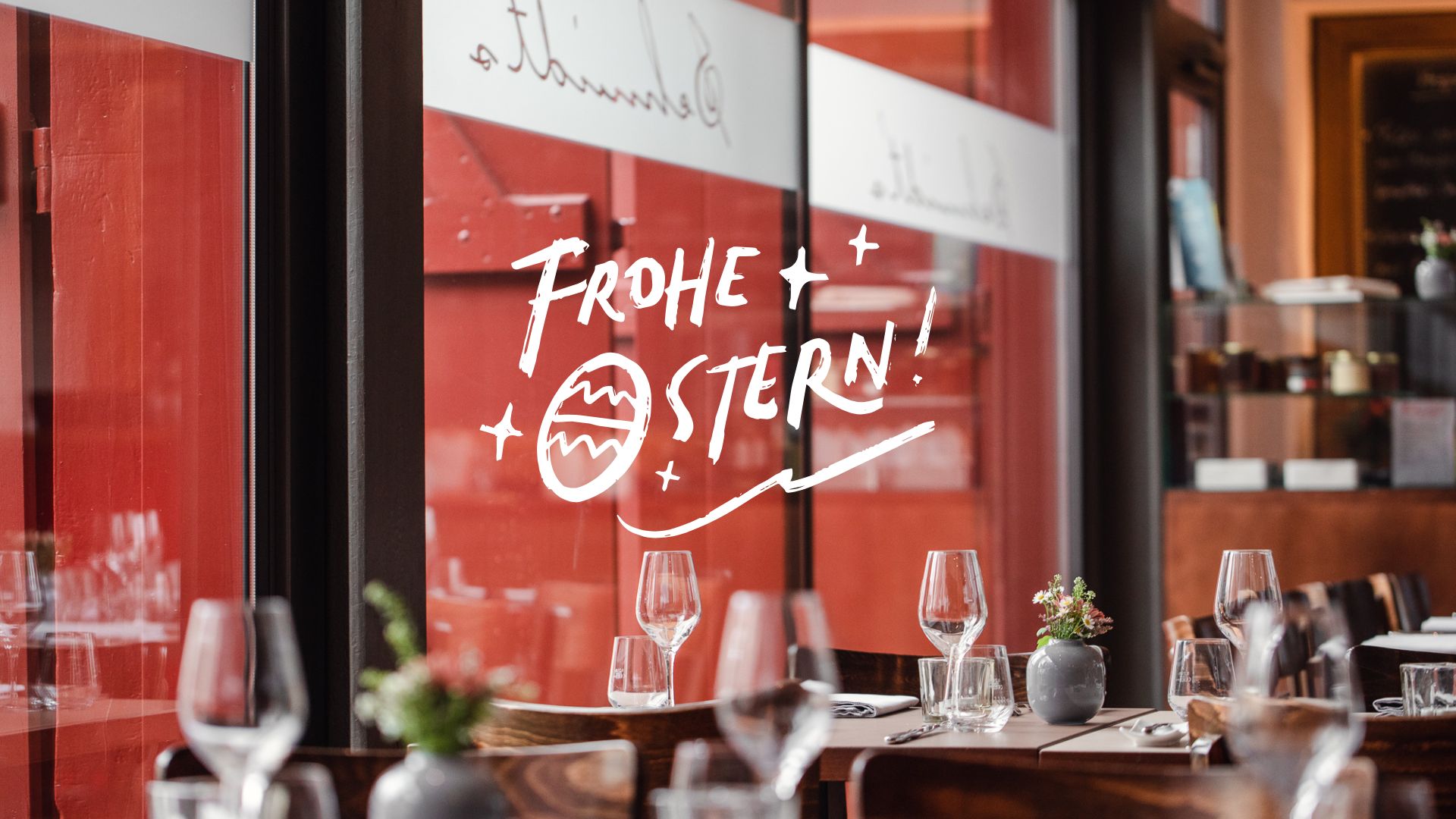 Auf der Fensterscheibe des Fine-Dining-Restaurants Schmidt’s in Dresden-Hellerau steht in festlichen, handgeschriebenen Buchstaben „Frohe Ostern!“. Die weiße Schrift mit kleinem Osterei-Motiv verleiht dem Innenraum eine saisonale, elegante Note. Im Vordergrund sind gedeckte Tische mit Gläsern und kleinen Blumenvasen zu sehen, die die warme Bistro-Atmosphäre des Restaurants unterstreichen.